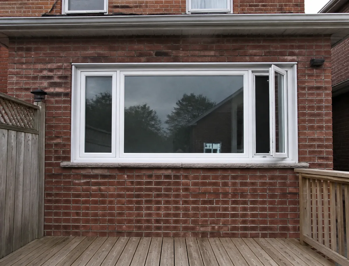 Back wall of a Toronto brick house with a white vinyl picture window and an opening casement on each side above a deck